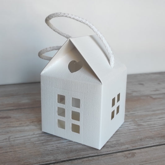 Scatola-Lanterna a forma di casetta in cartoncino fibra colore bianco. Misura 8 cm x 8 cm, H 12 cm  ed è completa di manico in corda.