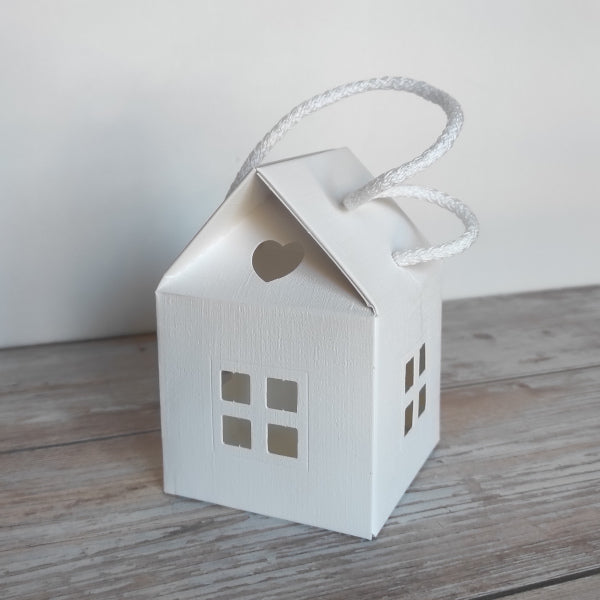 Scatola-Lanterna a forma di casetta in cartoncino fibra colore bianco. Misura 8 cm x 8 cm, H 12 cm  ed è completa di manico in corda.