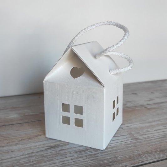 Scatola-Lanterna a forma di casetta in cartoncino fibra colore bianco. Misura 8 cm x 8 cm, H 12 cm  ed è completa di manico in corda.